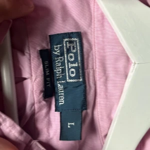Rosa skjorta från Polo Ralph Lauren - Snygg rosa skjorta från Polo Ralph Lauren i slim fit med klassisk button-down krage och broderad logga på bröstet. Skjortan är långärmad och har knappar framtill. Perfekt för dig som gillar stilrena och tidlösa plagg.