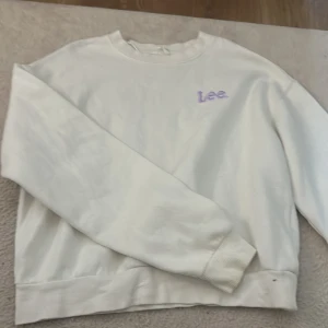 Vit sweatshirt från Lee - Säljer en clean vit sweatshirt från Lee med lila broderad logga på bröstet. Tröjan har rund hals och långa ärmar med ribbade muddar. Perfekt för dig som gillar enkel och stilren streetwear. Passar lika bra till jeans som till mjukisbyxor.