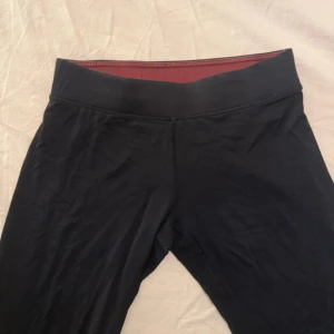 Svarta leggings från Newbody - Snygga svarta leggings från Newbody med bred midja och diskret logga på benet. De är stretchiga och sitter tight, perfekta för träning eller chill. Insidan har en mörkröd ton som ger en cool kontrast. Materialet känns mjukt och följsamt mot huden.