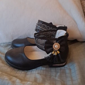 Svarta ballerinaskor med rosett och pärlor - Unika svarta ballerinaskor med rund tå. Skorna har breda meshband runt ankeln dekorerade med små pärlor och glittriga detaljer. På sidan sitter en stor rosett med pärlbroderi och hängande tofsar i guld och svart. Dragkedja bak för enkel påtagning.