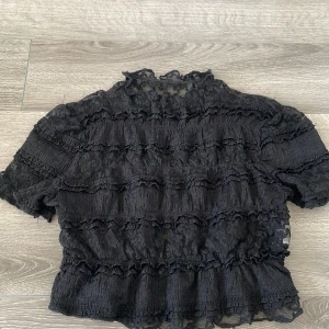 Svart blus med spets och volanger - Säljer en svart blus med korta ärmar, täckt av flera lager volanger och spetsdetaljer. Blusen har transparenta partier och en lätt rynkad struktur. Perfekt för dig som gillar att sticka ut med coola detaljer och unik design.