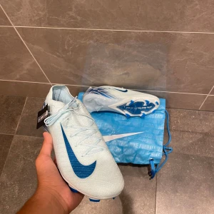 Nike Mercurial Air Zoom fotbollsskor - Nike Mercurial Air Zoom fotbollsskor i ljusblått med mörkblå detaljer och stor Swoosh på sidan. Skorna har snörning, platt sula med blå dobbar och en aerodynamisk form för maxad speed på planen. Ovandelen är i syntetmaterial och insidan har Mercurial-tryck. Levereras med blå Nike-påse.