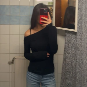 Svart oneshoulder långärmad topp - Snygg svart långärmad topp med oneshoulder-design som ger en cool och trendig look. Toppens passform är slim och materialet känns mjukt mot huden. Perfekt för dig som vill sticka ut med en enkel men stilren outfit.