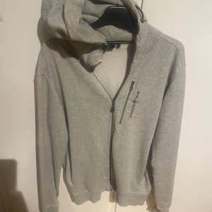 Den klassiska grå hoodien från Sail Racing  - Den klassiska grå hoodien från Sail Racing i storlek M. Tröjan har hel dragkedja framtill, en bröstficka med zip och broderad logga på bröstet. Mjuk insida och huva med snörning. Perfekt för chill dagar och riktigt snygg till jeans.
