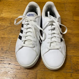 Vita Adidas sneakers med svarta ränder - Snygga vita sneakers från Adidas med klassiska svarta ränder på sidorna och blå Adidas-logga på plös och häl. Skorna har rund tå, platt sula och snörning framtill. Tillverkade i syntetmaterial med mjuk vadderad insida för extra komfort.