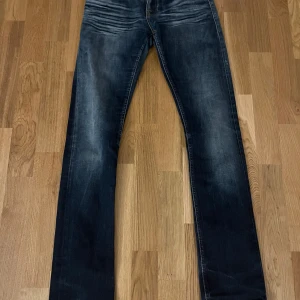Mörkblå Nudie Jeans Super Slim - Snygga mörkblå jeans från Nudie Jeans i modellen Super Slim. Jeansen har en smal passform med klassisk femficksdesign och coola slitningar framtill. Materialet är jeans med orangea kontrastsömmar som ger en schysst detalj. Perfekt för dig som gillar en tight look.