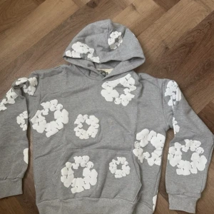 Grå hoodie med vita blommor från Denim Tears - Säljer en grå hoodie från Denim Tears med stora vita blommor tryckta över hela tröjan. Tröjan har huva, magficka och ribbade muddar vid ärmslut och nederkant. Perfekt för dig som gillar streetwear med unik design och vill sticka ut.