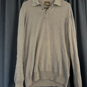 Grå långärmad piké  från Oscar Jacobson - Snygg grå pikétröja från Oscar Jacobson i storlek S. Tröjan har klassisk krage, tre knappar vid halsen och långa ärmar. Materialet känns mjukt och skönt mot huden, perfekt för dig som gillar stilrena och enkla plagg.