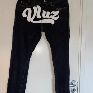 Svarta jeans från Vluz med tryck - Unika svarta jeans från Vluz med stort vitt tryck framtill och broderade detaljer på bakfickorna. Snygga kontrastsömmar och klassisk femficksdesign. Materialet är robust denim och passformen är rak. Perfekt för dig som vill sticka ut med cool streetstil.