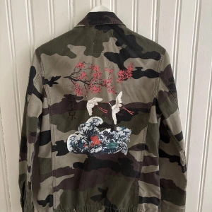 Premium collection Bershka jacka med tryck - Säljer en grön camouflage jacka med snygga detaljer på ryggen. Jackan har klassisk krage, knappar framtill och flera fickor. Perfekt för dig som vill ha en cool och avslappnad stil. Från Bershka men säljs ej längre