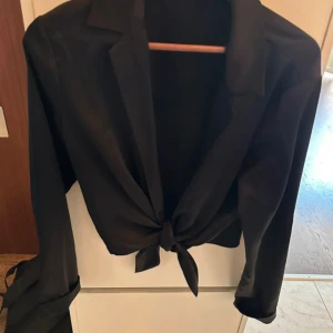 Svart croppad skjorta från Bik Bok - Svart skjorta från Bik Bok i croppad modell med knyt framtill. Skjortan har klassisk krage och långa ärmar.  Perfekt för dig som gillar stilrena och trendiga plagg.