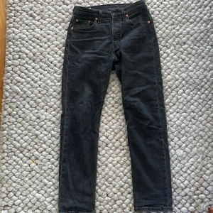 Levi's 501 svarta jeans W25 L28 - Svarta Levi's 501 jeans med klassisk rak passform och fem fickor. Jeansen har knappgylf och detaljer i kopparfärgad metall. Materialet är robust denim i bomull och färgen är mörk svart. Perfekt för dig som gillar tidlös och clean stil.