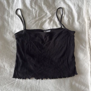 Svart ribbad croppad linnetopp - Svart croppad linnetopp med smala axelband och ribbad struktur. Toppen har en vågig kant nedtill som ger en chill vibe. Perfekt att matcha med jeans eller kjol för en enkel och snygg look.
