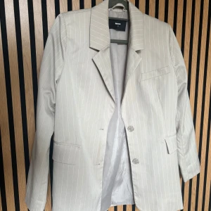 Beige randig kavaj från Bik Bok - Snygg beige kavaj från Bik Bok med smala vita ränder. Klassisk krage, två knappar framtill och fickor med lock. Perfekt för dig som gillar stilrena och trendiga plagg. Passar till både jeans och kostymbyxor.