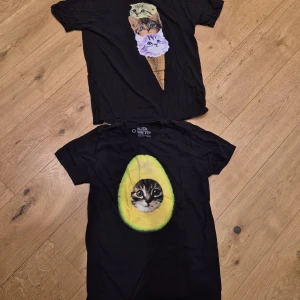 Svarta t-shirts med kattmotiv st M - Två svarta t-shirts i bomull med unika kattmotiv. Ena har en katt i en avokado, den andra har tre kattansikten staplade som en glass. Båda har rund hals och korta ärmar. Perfekt för dig som gillar roliga och annorlunda prints. Storlek M