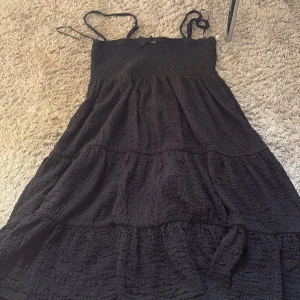Svart långklänning från Gina Tricot - Säljer en svart långklänning från Gina Tricot i storlek M. Klänningen har smock upptill, tunna axelband och en liten rosett framtill. Materialet är mjukt och har en skrynklig struktur med volanger i kjolen för extra flow. Perfekt för dig som gillar enkel men snygg stil.