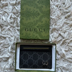 Svart designer plånbok i läder - Snygg svart plånbok (får ej skriva märket i beskrivningen då annonsen tas bort med det står på lådorna på bilderna)  med klassiskt GG-mönster i grått. Plånboken har flera kortfack och en metallfärgad GG-logga på framsidan. Kommer med original ask, dustbag och äkthetskort. Perfekt för dig som vill ha en stilren och lyxig accessoar. Plånboken är sprillans ny och aldrig använd i ett väldigt mjukt läder. Alla lådor och kort medföljer vid köp!