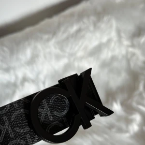 Svart Calvin Klein bälte  - Snyggt svart bälte från Calvin Klein med coolt CK-logomönster över hela bandet. Bältet har en stor, rund CK-spänne i svart metall som ger en modern vibe. Tillverkat i skinn med en stilren och trendig look som lyfter din outfit.