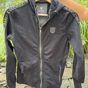 Svart vindjacka från Maya Delorez XS - Svart vindjacka från Maya Delorez med huva och dragkedja framtill. Snygga logodetaljer på bröstet och längs axlarna. Jackan har två fickor med dragkedja och justerbar snörning i huvan. Perfekt för dig som gillar sportig och clean stil.