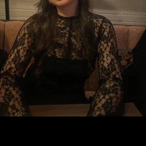 Svart spetsblus med lång ärm - Supersnygg svart blus i transparent spets med blommigt mönster. Långärmad och figurnära passform, perfekt att styla med ett linne under eller som den är för en edgy look. Hög hals och detaljerad spets gör den extra cool.