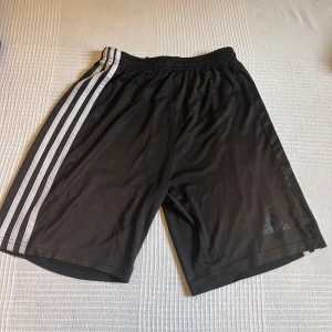 Svarta Adidas shorts med vita ränder - Svarta sportiga shorts från Adidas med klassiska vita ränder längs sidorna och Adidas-logga framtill i storlek M. De har elastisk midja och är gjorda i ett lätt och skönt syntetmaterial, perfekta för träning eller chill. Snygg loose passform och stilren design.