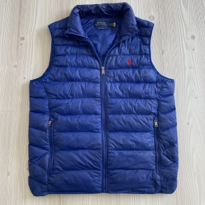Blå dunväst från Polo Ralph Lauren - Snygg blå dunväst från Polo Ralph Lauren med horisontella quiltade paneler och dragkedja framtill. Västen har två fickor med dragkedja och en klassisk ståkrage. Ikonisk röd Polo-logga broderad på bröstet. Perfekt för lager-på-lager och chill stil. Väldigt unik färg som inte tillverkas längre. Ända som säljs på den begagnade marknaden. Skicket är 10/10, finns inga tecken på användning. Storlek S passar M också. Nypris 3599:- mitt pris 1299:- frågor? Bara att höra av sig!