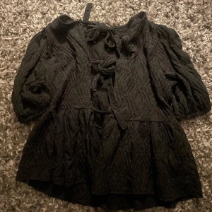 Svart blus med knyt från Gina Tricot - Supersnygg svart blus från Gina Tricot i storlek S. Blusen har ett coolt strukturmönster, puffiga trekvartsärmar och flera knyt framtill som ger en unik vibe. Den är lite volangig nedtill och har en loose passform. Perfekt för dig som gillar detaljer och vill sticka ut.