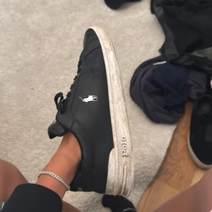 Svarta Polo Ralph Lauren sneakers - Svarta sneakers från Polo Ralph Lauren med vit logga på sidan och Polo-tryck på plösen och sulan. Skorna har rund tå, snörning och en vit platt sula i syntet. Stilren design med klassisk streetkänsla, perfekt för dig som gillar enkel men snygg look.