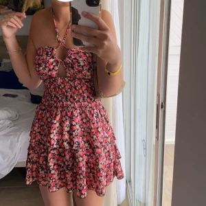 Blommig kort sommarklänning - Supersöt kort klänning med blommigt mönster i rosa, rött och svart. Klänningen har smockad midja, volangig kjol och halterneck med snygg cut-out framtill. Perfekt för varma dagar och ger en riktigt fräsch vibe.
