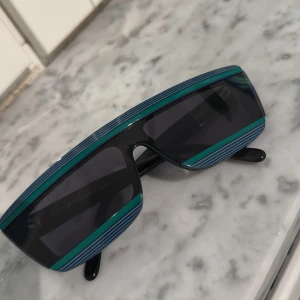 Randiga solglasögon Courrèges Paris - Säljer ett par unika solglasögon från Courrèges Paris, handgjorda i Frankrike. Modellen har en futuristisk, rektangulär form med svarta och mörkgröna bågar och coola blå ränder längs ovansidan och nederkanten. Perfekt statement-piece för dig som gillar att sticka ut.