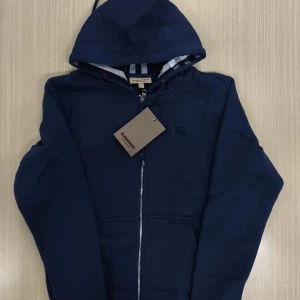 Marinblå hoodie från Burberry - Snygg marinblå hoodie från Burberry med dragkedja framtill och huva. Jackan har klassiska Burberry-detaljer på insidan av huvan och en diskret logga på bröstet. Perfekt för dig som gillar stilrena och exklusiva plagg.