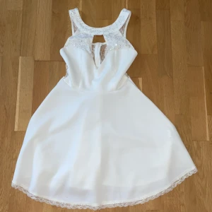 Vit kort klänning med paljetter - Supersöt vit klänning med glittriga paljetter upptill och transparent mesh vid axlarna. Klänningen har en snygg öppning fram och bak, samt spetskant längst ner på kjolen. Passar dig som gillar en feminin och festlig stil med lite extra bling.