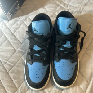 Nike Air Jordan 1 Low blå/svarta - Säljer ett par Nike Air Jordan 1 Low i blått och svart med klassisk Jumpman-logga på tungan och Air Jordan-vinge på hälen. Skorna har snörning, perforerad tå och platt sula. Snygg kontrast mellan blått och svart skinn, perfekt för dig som gillar streetstyle.