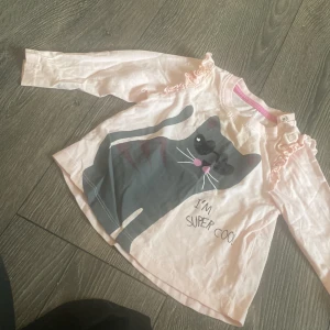 Ljusrosa långärmad tröja med katt - Söt ljusrosa långärmad tröja från Petit Litet med tryck av en cool katt med solglasögon och texten 'I'm super cool'. Tröjan har volangdetaljer på axlarna och praktiska knappar vid halsen. Tillverkad i mjuk bomull, perfekt för dig som gillar gulliga prints.