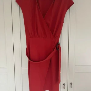 Röd omlottklänning från NA-KD - Snygg röd omlottklänning från NA-KD med v-ringning och korta ärmar. Klänningen har ett brett bälte med svart ringdetalj i midjan som ger en cool touch. Modellen är kort och har en feminin passform som framhäver figuren. Perfekt för dig som gillar stilrena och trendiga plagg.