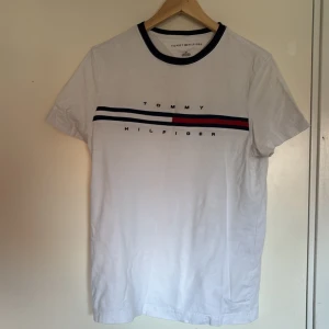 Vit Tommy Hilfiger t-shirt med ränder - Vit t-shirt från Tommy Hilfiger med klassisk rund hals och korta ärmar. Framsidan har tryck med blå och röd rand samt logotypen. Halsringningen är mörkblå och materialet är mjuk bomull. Perfekt för en clean och stilren look.