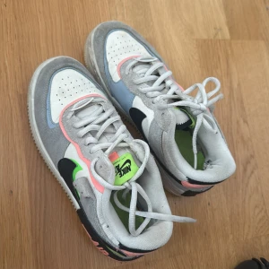 Nike Air Force 1 sneakers med färgdetaljer - Nike Air Force 1 sneakers med en mix av grå mocka, vit textil och detaljer i neon-grönt, ljusblått och rosa. Snygg svart swoosh på sidan och AIR-text bak. Skorna har snörning och platt sula, perfekt för dig som gillar streetstyle och färg.