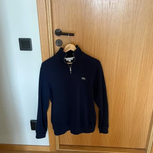 Marinblå half zip tröja från Lacoste - Snygg marinblå tröja från Lacoste med klassisk krokodillogga på bröstet. Half zip-dragkedja och hög krage ger en clean look. Tillverkad i mjuk bomull, perfekt för chill dagar. Långärmad och normal passform. Tröjan är i stort sätt oanvänd och därav nästan i nyskick