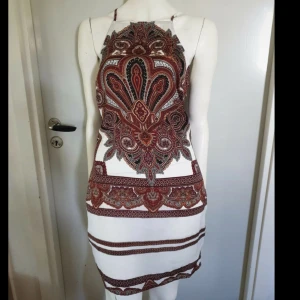 Vit kortklänning med paisleymönster - Snygg vit kortklänning med tunna axelband och paisleyinspirerat mönster i brunt, svart och rött. Klänningen har en rak passform och är ärmlös, med coola detaljer längs sidorna. Skulle passa storlek 34-36, fast det är en 38a