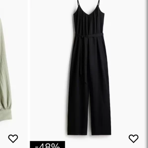 Svart byxdress med smala axelband - Snygg svart byxdress med smala axelband och v-ringad hals. Modellen har ett bälte i midjan och vida ben som ger en chill och trendig look. Perfekt för dig som gillar enkel stil med en twist. Materialet ser ut att vara mjukt och följsamt.