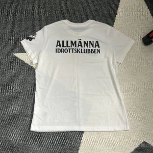 Vit Nike AIK t-shirt Dri-FIT L - Vit t-shirt från Nike med AIK-logga på ärmen och stor svart swoosh på bröstet. Baksidan har trycket 'ALLMÄNNA IDROTTSKLUBBEN' i svart. Tillverkad i mjukt Dri-FIT material som andas, perfekt för fotboll eller träning. Klassisk rund hals och korta ärmar.