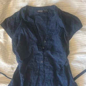 Mörkblå blus med knyt - Snygg mörkblå blus från Esmara med korta ärmar och diskret broderat mönster. Blusen har v-ringning med knappar framtill och ett knytband i midjan som ger en schysst siluett. Perfekt för dig som gillar enkel men stilren design. Storlek 38 men skulle säga att den även passar mindre då man själv kan välja hur mycket man drar åt!