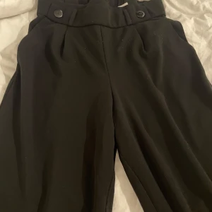 Svarta kostymbyxor med knappar - Snygga svarta kostymbyxor med bred resår i midjan och två dekorativa knappar framtill. Byxorna har en rak och lite loose passform med klassisk look. Perfekta att styla med en cool t-shirt eller hoodie för en chill vibe.