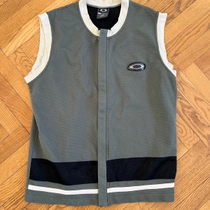 Khaki meshväst från Oakley M - Sportig khaki meshväst från Oakley med vita detaljer vid ärmslut och krage. Västen har svart och vit rand längst ner och Oakley-logga på bröstet. Från 90-talet. Perfekt för dig som gillar streetwear och sportig stil. Lätt och luftig design med stängning framtill.