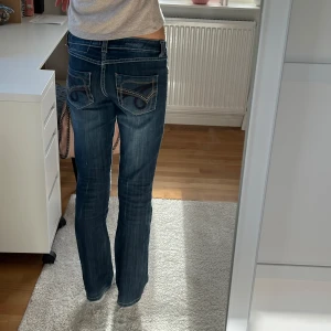 Blå bootcut jeans med broderade fickor - Snygga blå jeans med bootcut passform och låg midja. Jeansen har coola broderade detaljer på bakfickorna och kontrastsömmar som ger en extra touch. Materialet är klassisk jeans och färgen är mörkblå med lätt slitning. 