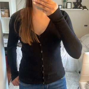 Svart ribbad kofta med v-ringning - Säljer en svart, ribbad kofta med v-ringad hals och långa ärmar. Koftan har en rad svarta knappar framtill och sitter snyggt med en slim passform. Perfekt att slänga över en t-shirt eller bära som den är. Materialet är mjukt och stickat. Jag bär oftast S, och den är lite för tajt på mig. Skulle ändå säga att den passar xs och s, om man vill ha den lite tajt!😇