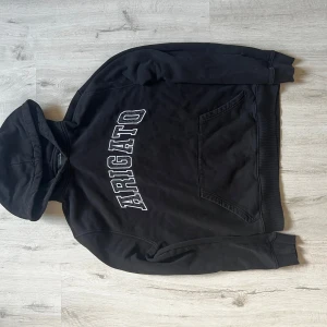 Svart hoodie Axel Arigato med tryck - Svart hoodie från Axel Arigato med stor vit text på bröstet. Tröjan har huva, känguruficka och ribbade muddar. Tillverkad i 100% ekologisk bomull, perfekt för chill dagar. Snygg och enkel design som funkar till jeans eller joggers.