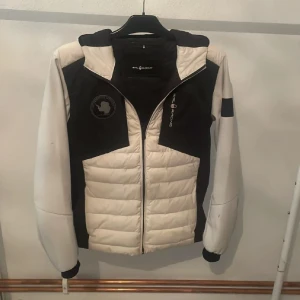 Vit och svart pufferjacka Sail Racing - Snygg pufferjacka från Sail Racing i vit och svart med broderad Sail Antarctica Expedition-logga på bröstet. Jackan har huva, dragkedja och quiltad front. Perfekt för dig som gillar sportig stil och vill ha en jacka som sticker ut.