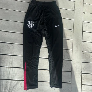 Svarta Nike FC Barcelona träningsbyxor S - Svarta träningsbyxor från Nike med FC Barcelona-logga på ena benet och vit Nike-swoosh. Byxorna har elastisk midja, smal passform och färgdetaljer i rött och blått vid bensluten. Tillverkade i polyester, perfekta för fotboll eller träning.
