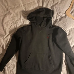 Svart hoodie från Polo Ralph Lauren - Svart hoodie från Polo Ralph Lauren med klassisk röd broderad logga på bröstet. Tröjan har huva, magficka och långa ärmar. Kontakta vid funderingar. Priset kan diskuteras vid snabb affär.